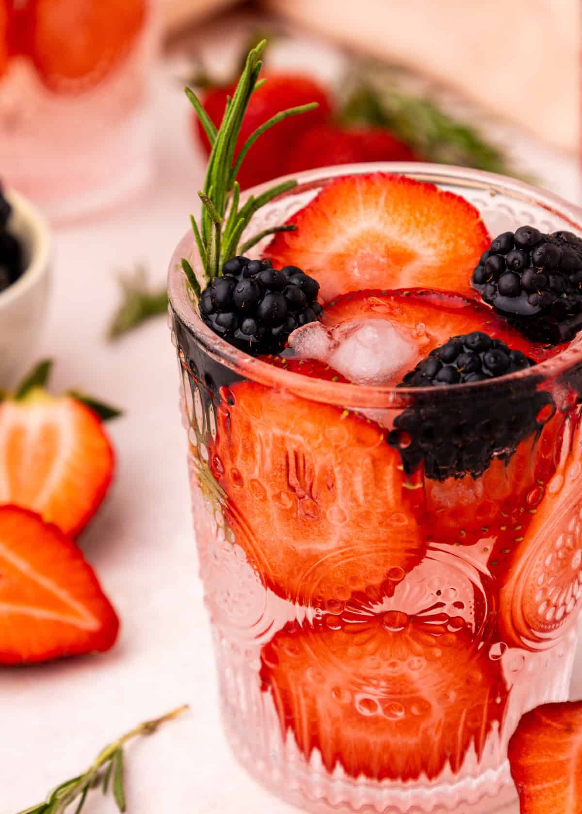 A glass of strawberry water with sliced strawberries, blackberries, and a sprig of rosemary.