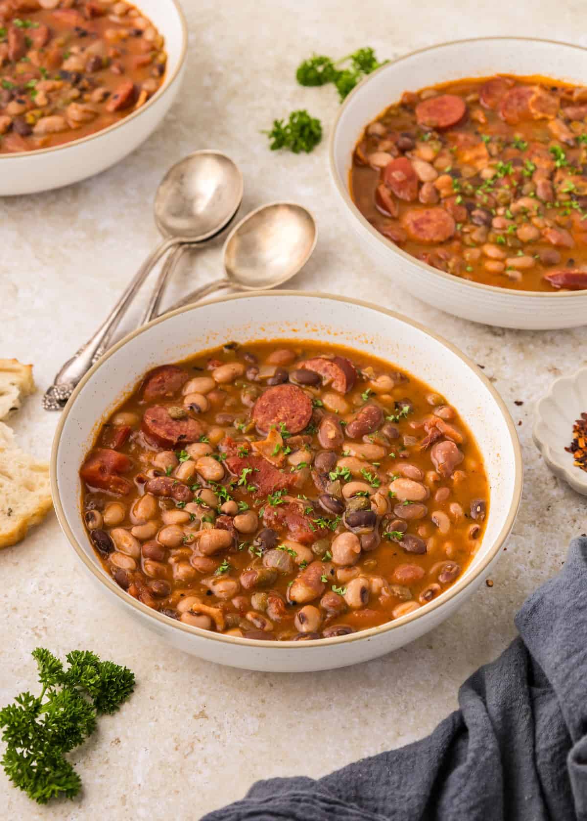 A bowl of bean soup with sausage, garnished with parsley, served with bread and spoons.