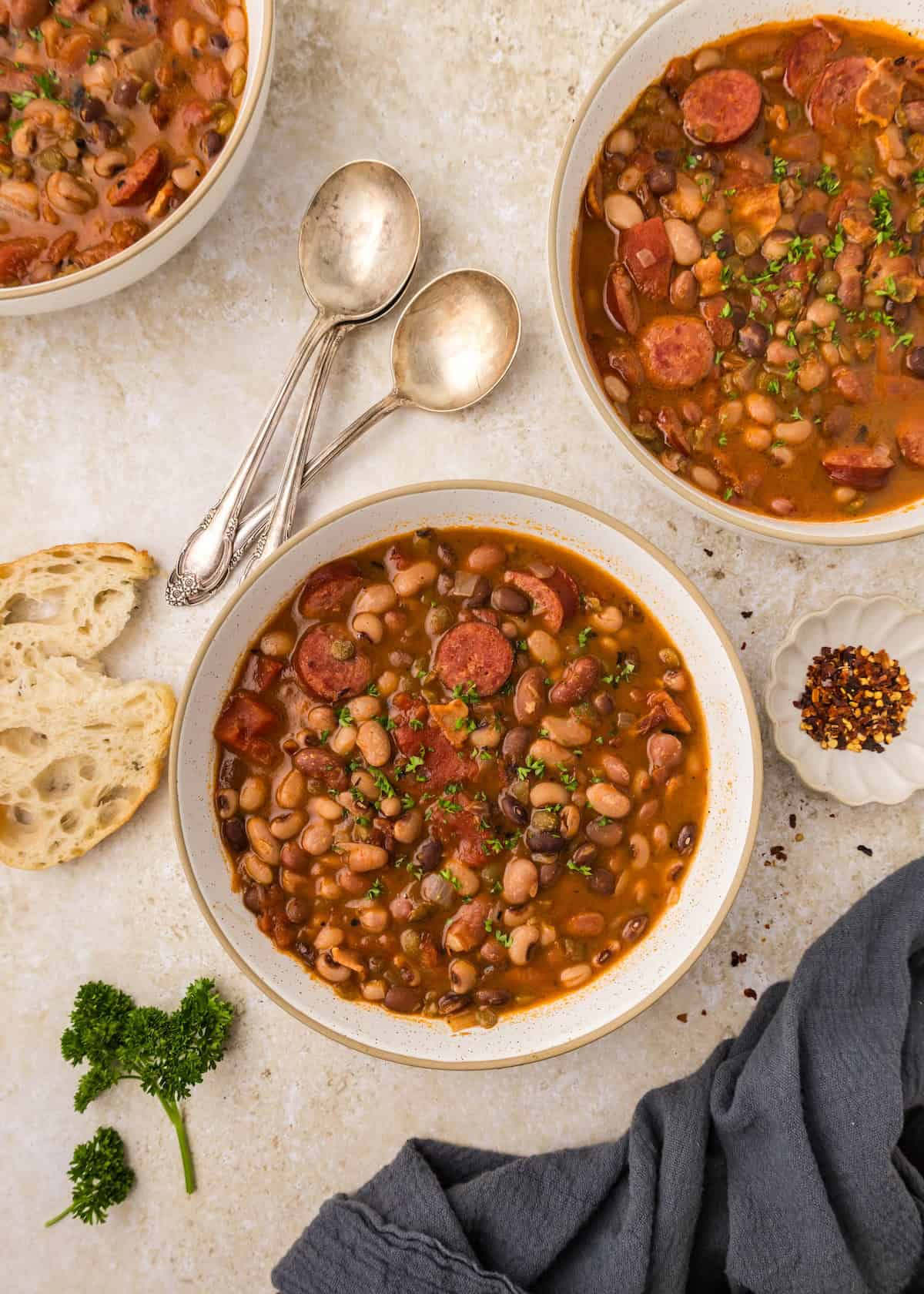Bowls of hearty bean soup with sausage, bread, parsley, spoons, and red pepper flakes on the side.