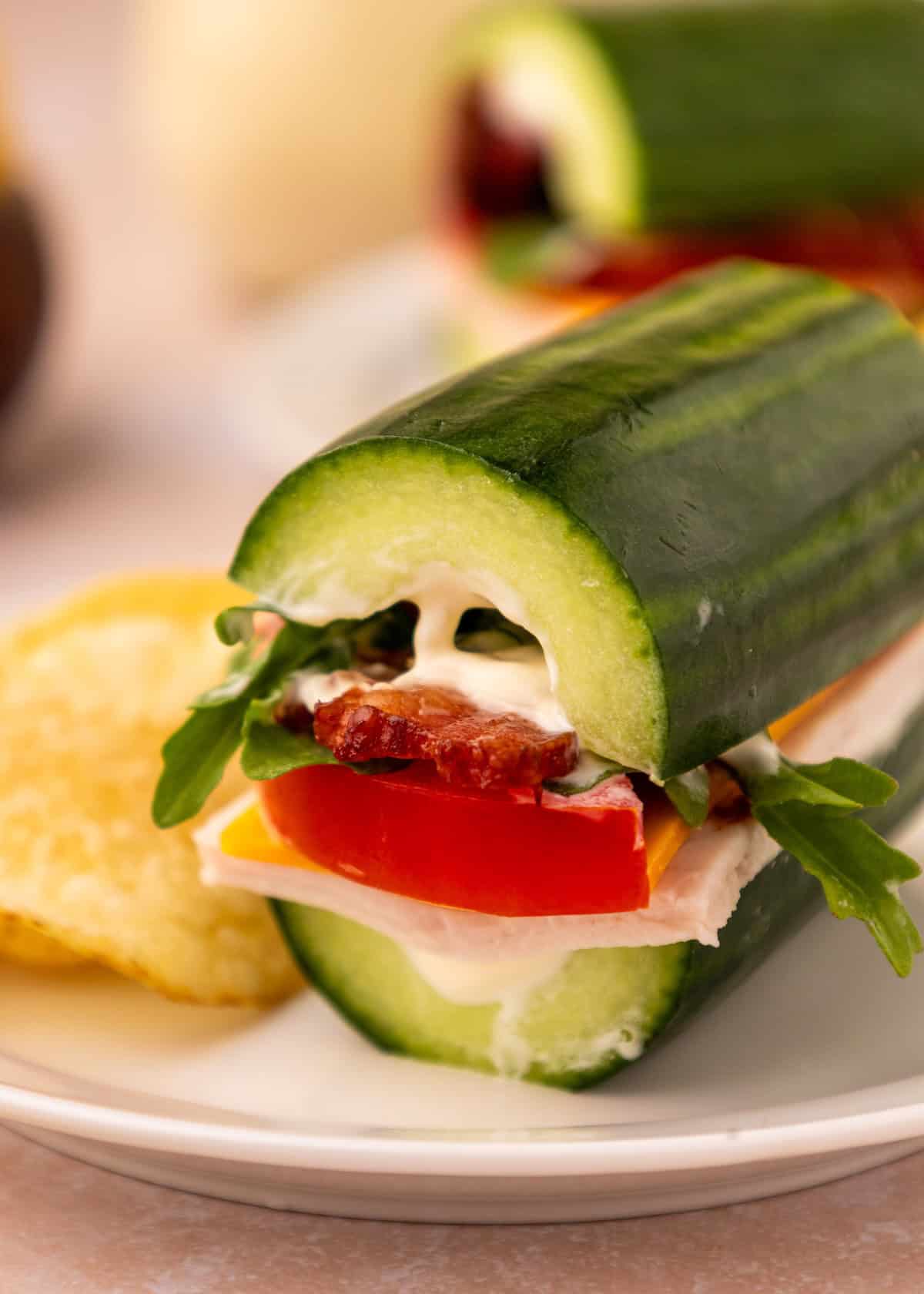 Cucumber subs with turkey, cheese, tomato, bacon, greens, and mayo on a white plate.