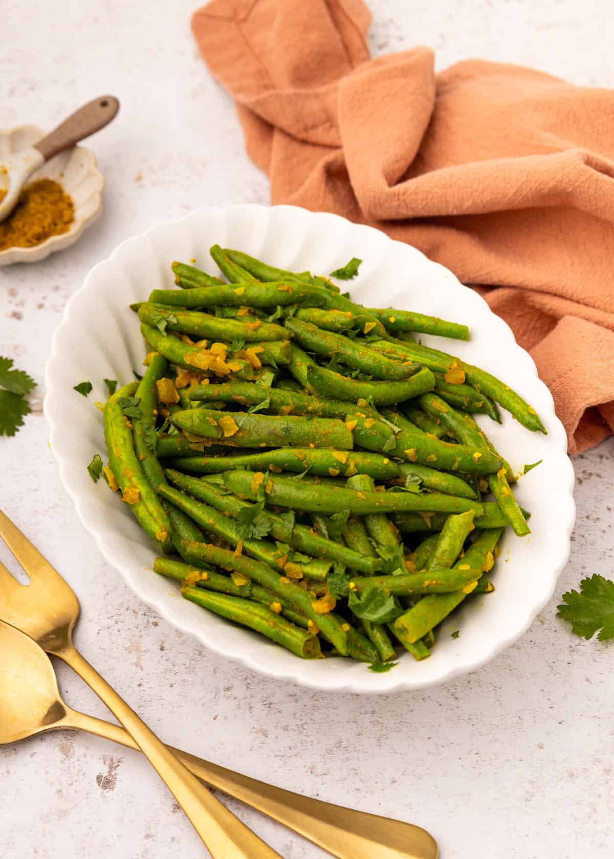Curried green beans with spices served in a white oval dish, gold utensils, and napkin nearby.