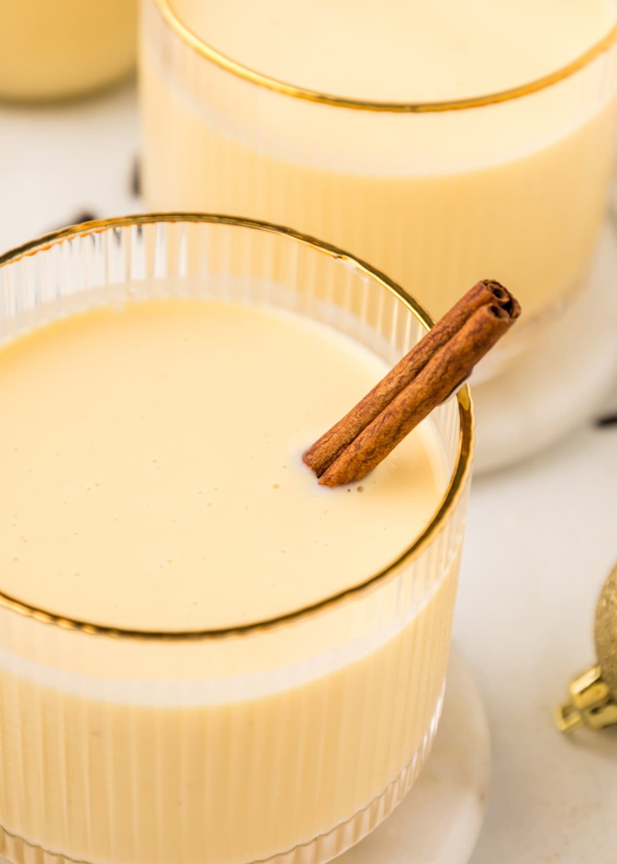 A glass of homemade eggnog with a cinnamon stick, rimmed with gold, on a white surface.