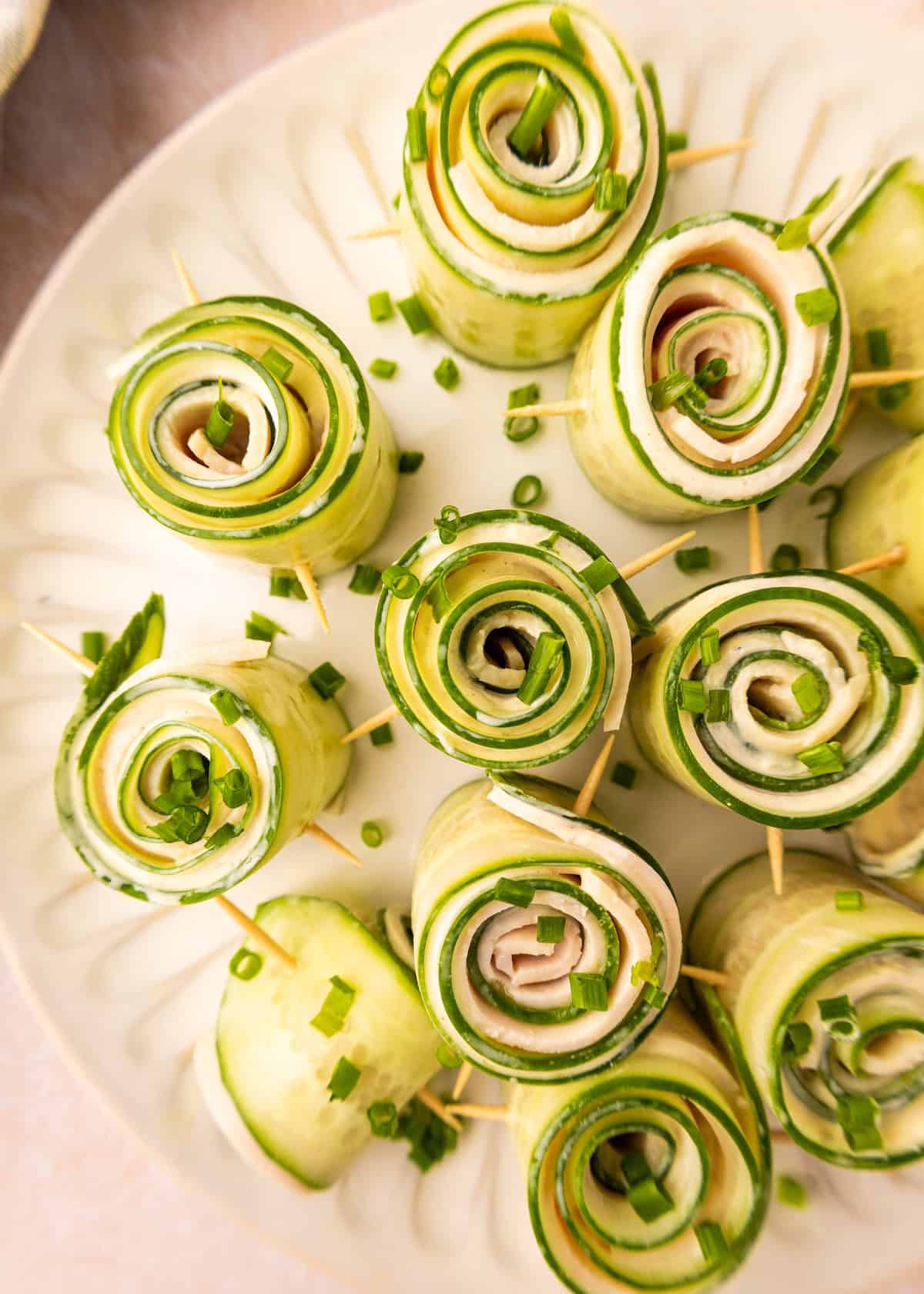 Cucumber rolls and ham roll-ups with toothpicks, garnished with chives on a white plate.