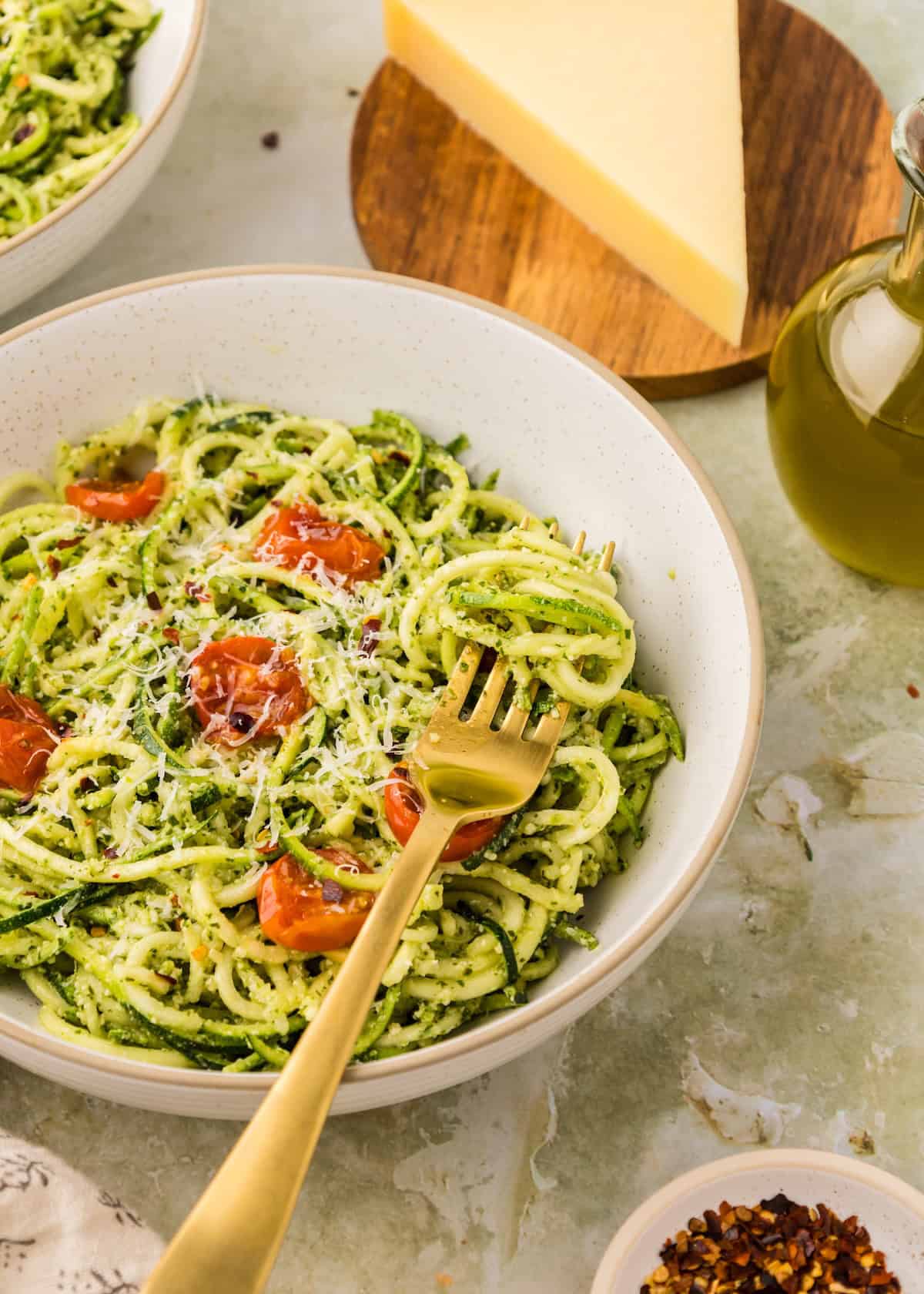 Bowl of pesto zucchini noodles with cherry tomatoes, grated cheese, and a gold fork by cheese wedge.