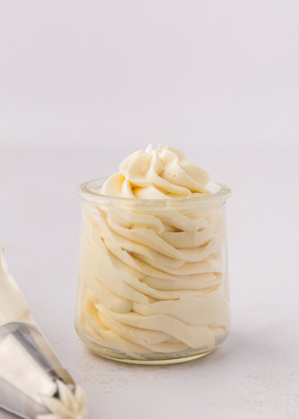 A small glass jar filled with swirled cream cheese frosting next to a piping bag.