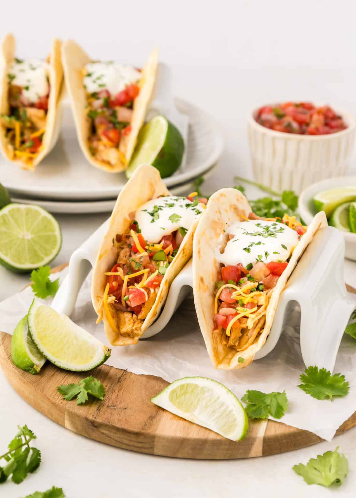 Two crockpot shredded chicken tacos with toppings and lime wedges on a wooden board.