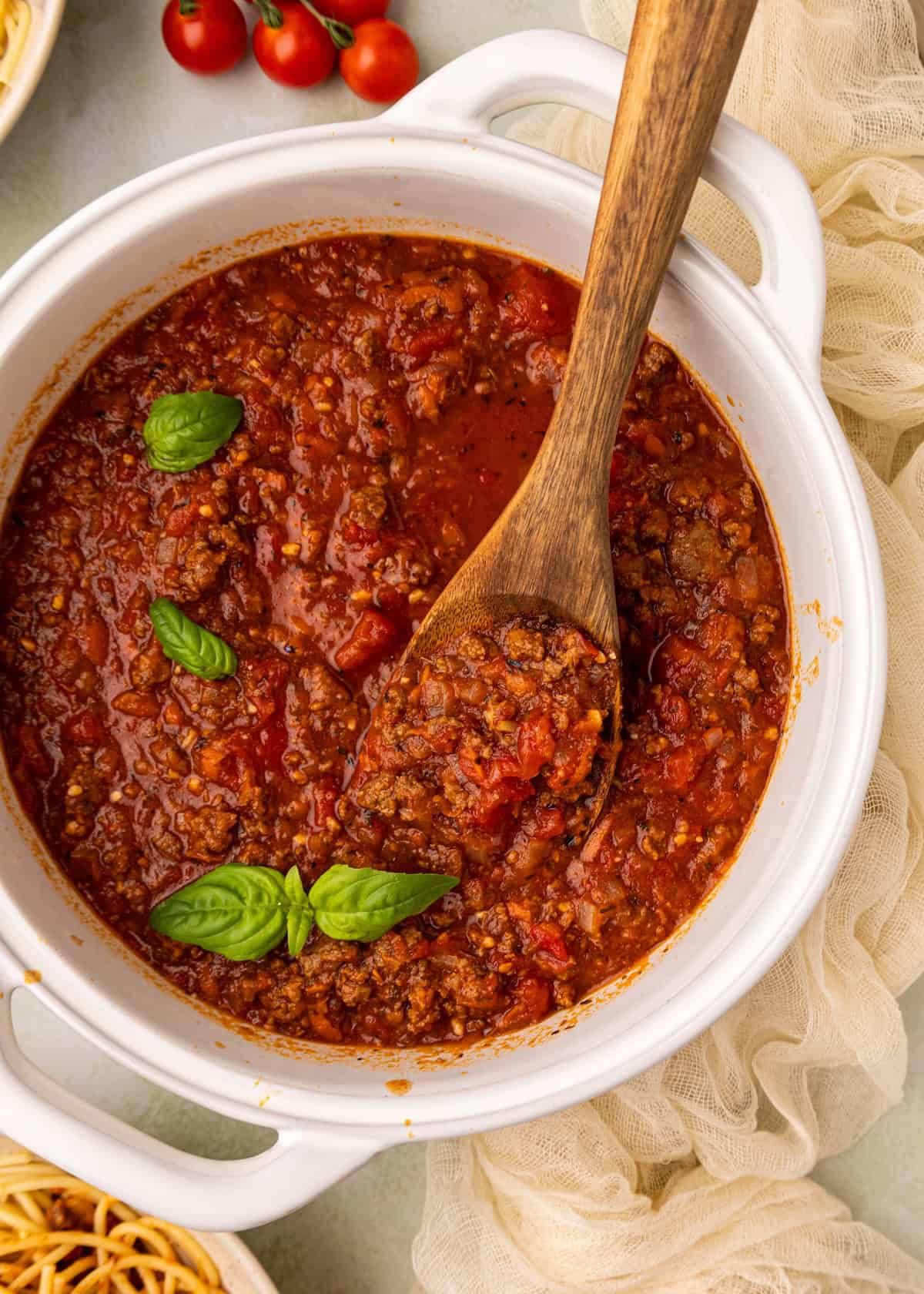 Homemade spaghetti sauce in a white pot with basil leaves and a wooden spoon.