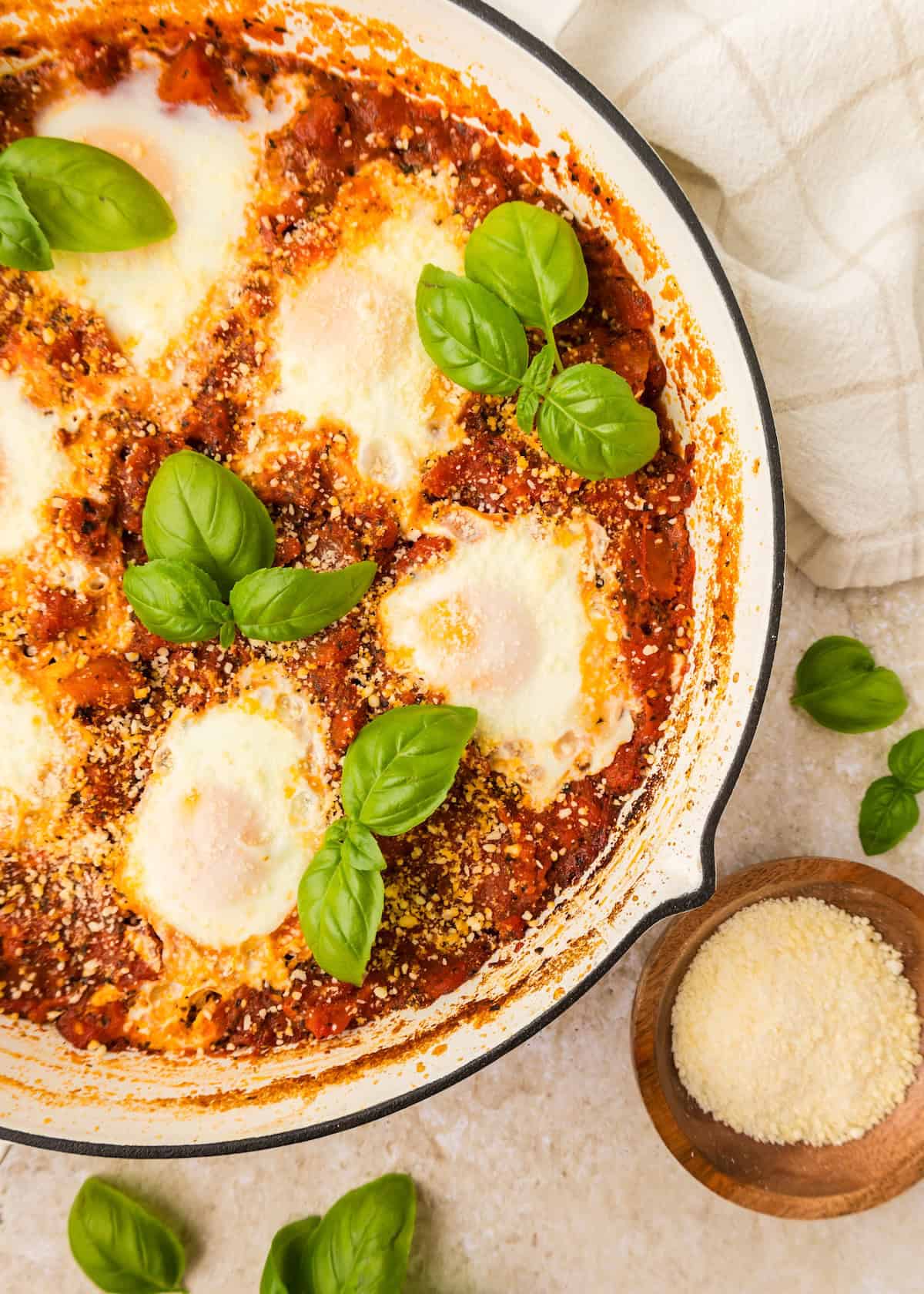 Eggs in purgatory baked in tomato sauce, topped with basil and cheese in a white skillet.