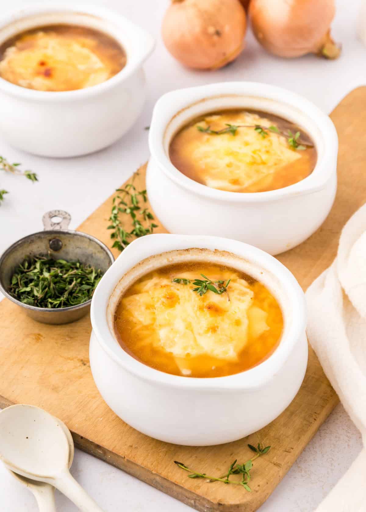 Three bowls of french onion beef soup with melted cheese, herbs, and onions on a wooden board.