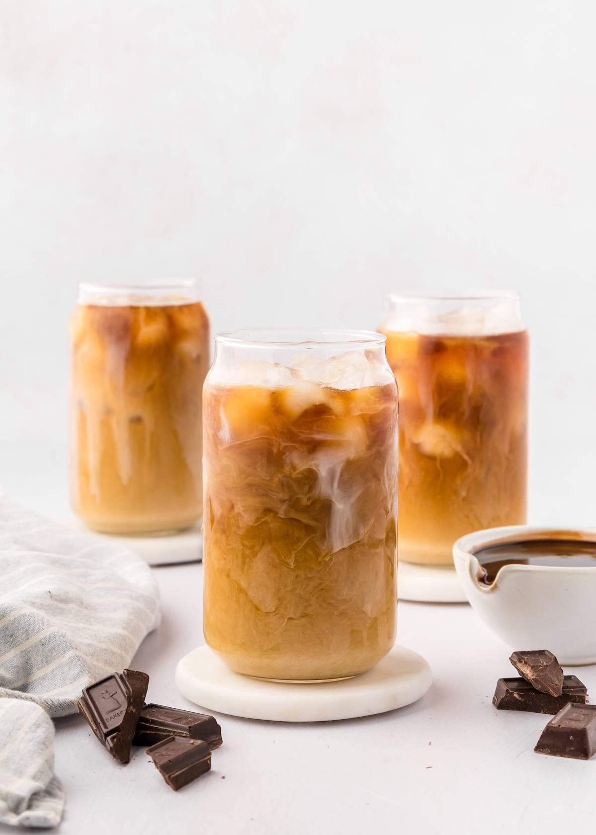 Three glasses of iced mocha with milk, surrounded by chocolate pieces and a small bowl of syrup.