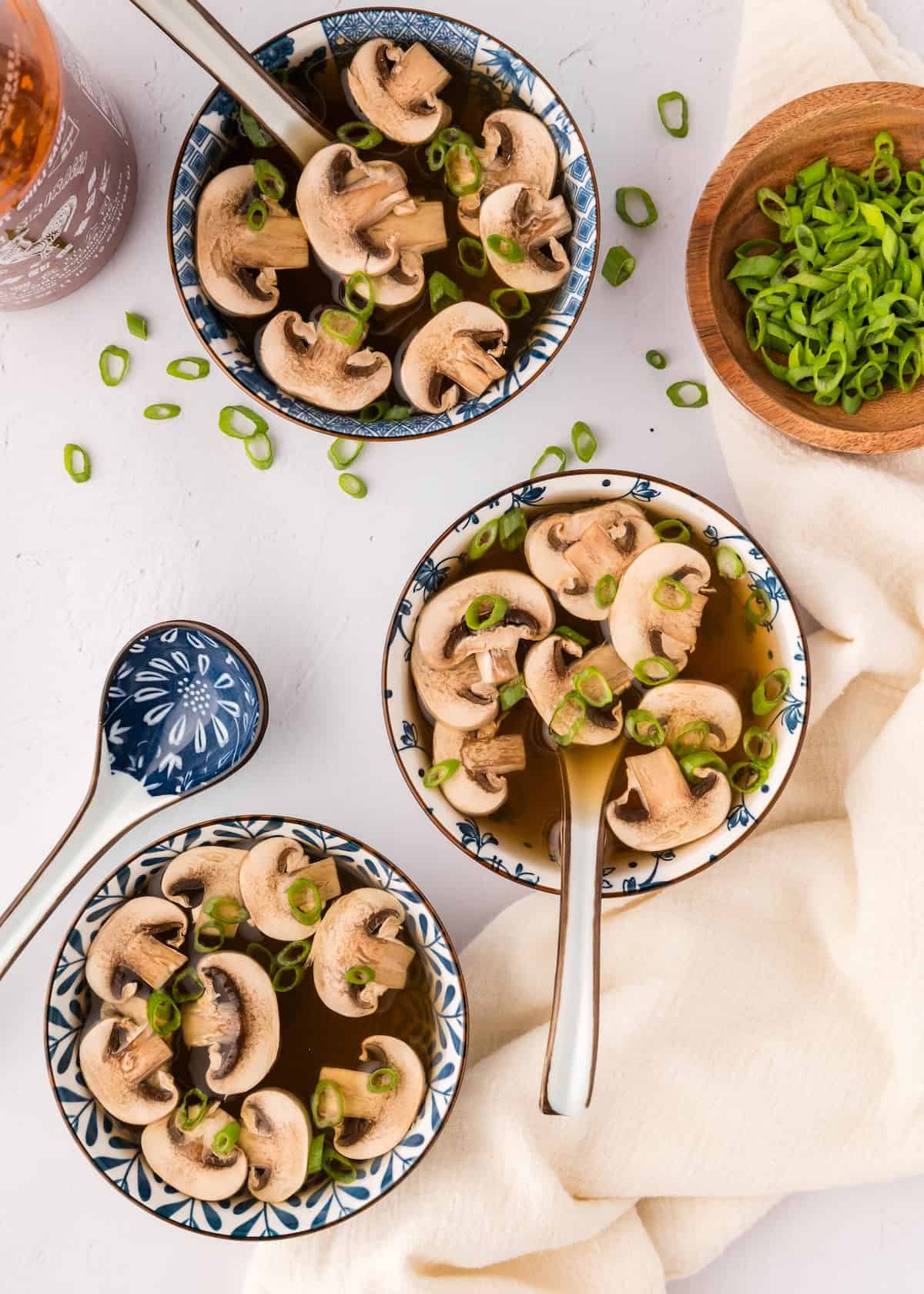 Three bowls of Japanese clear onion soup topped with sliced green onions, beside more onions.