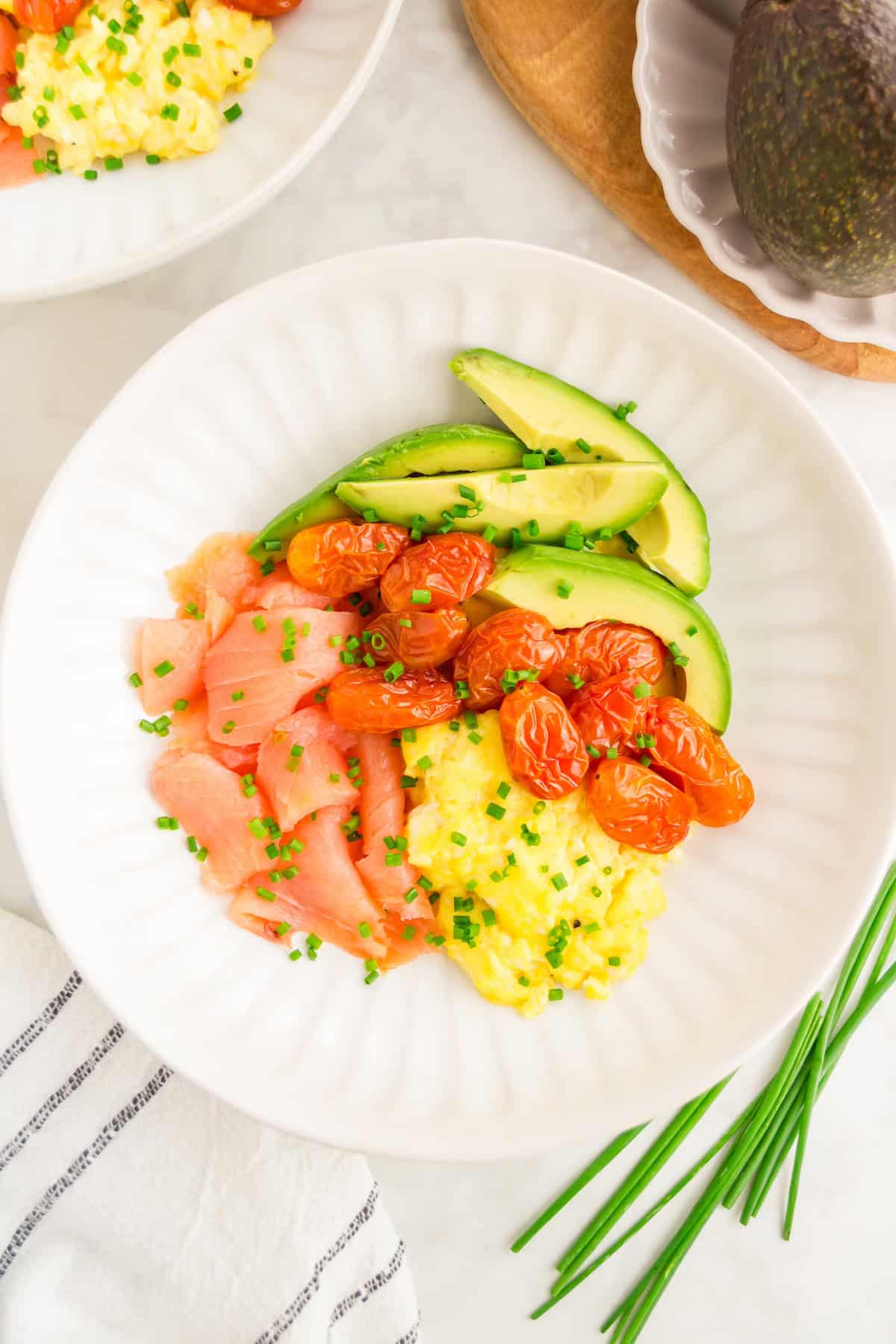 White plate with a smoked salmon breakfast bowl of eggs, tomatoes, avocado, and chives.