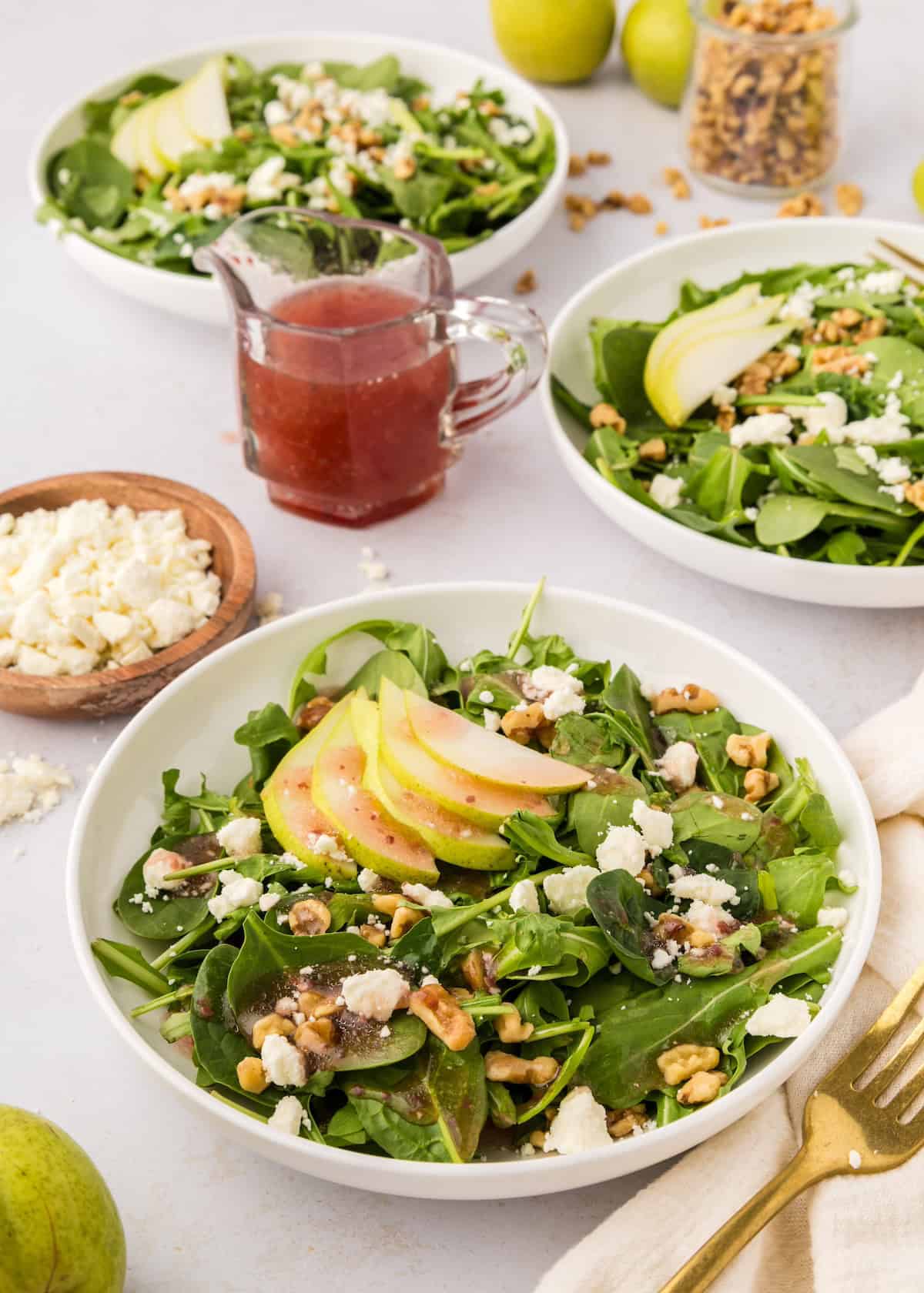 Three bowls of walnut pear salad with spinach, apple slices, feta, and a jar of red vinaigrette.
