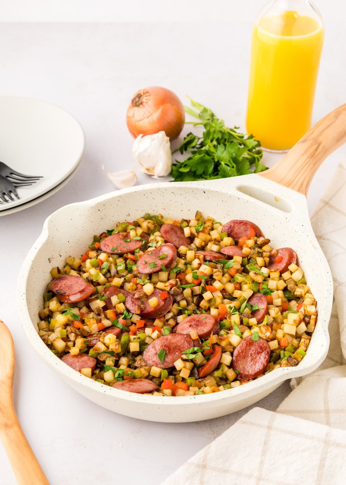 A skillet of smoked sausage hash with fresh herbs, onion, and a glass of orange juice.