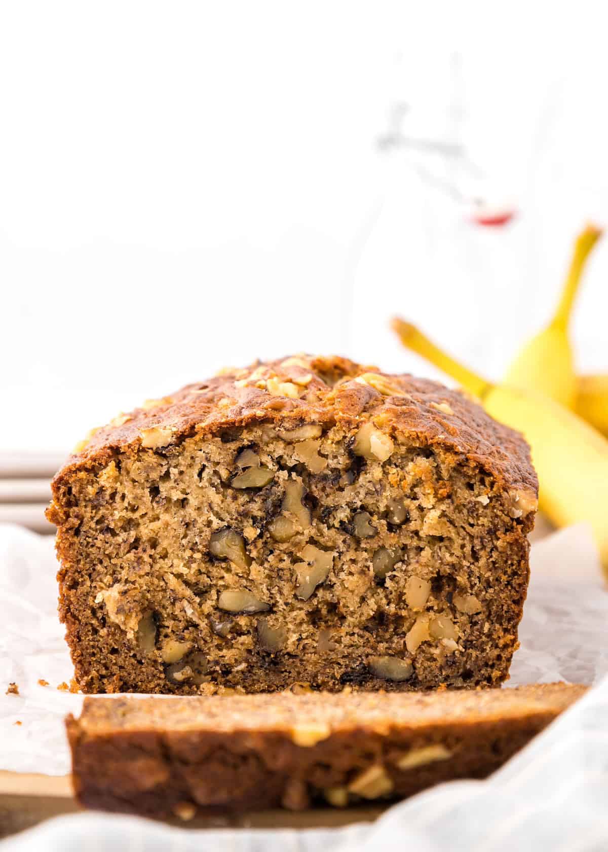 A sliced loaf of Einkorn Banana Bread with walnuts on parchment, bananas in the background.