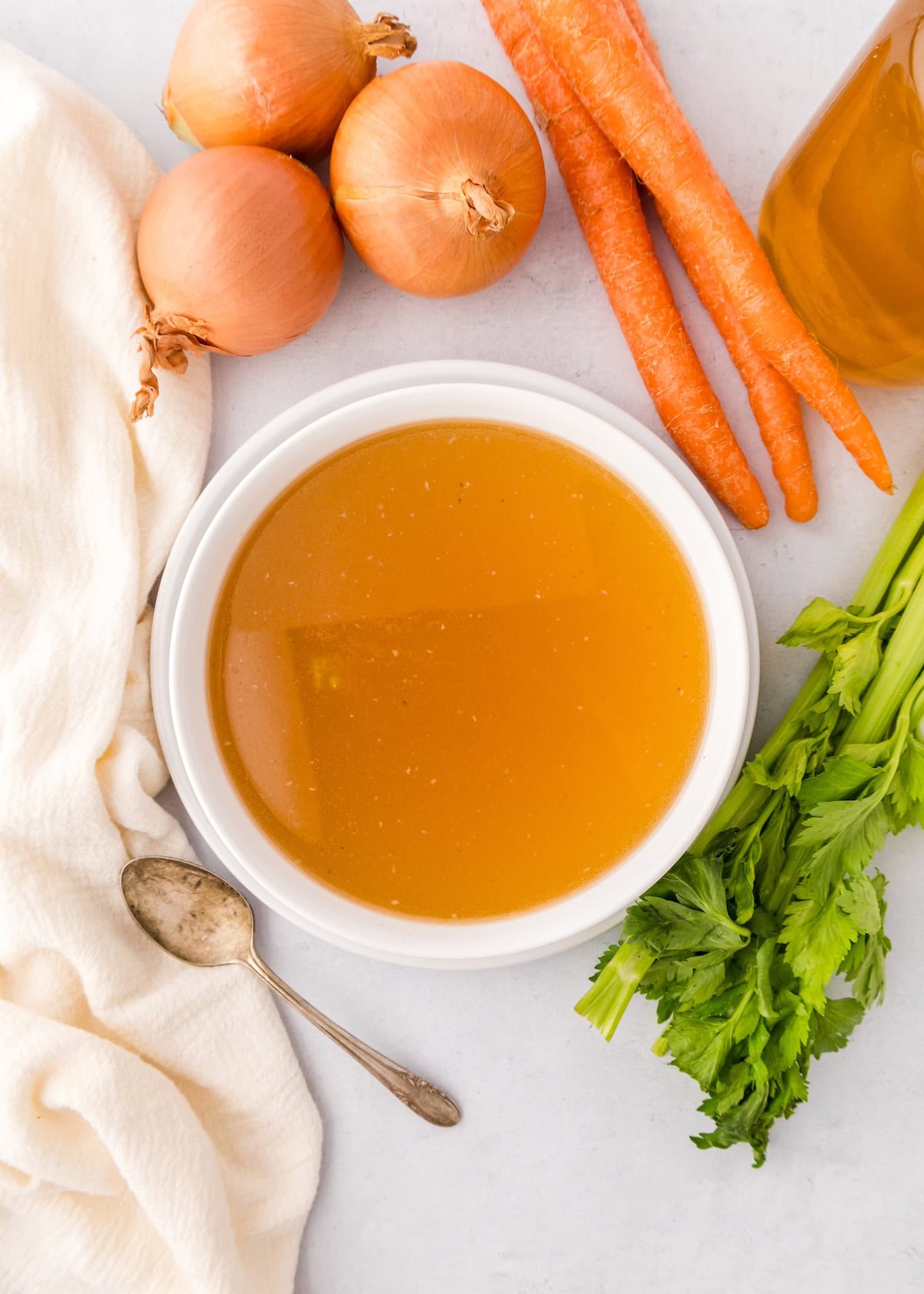 Bowl of Chicken Bone Broth with onions, carrots, celery, a spoon, and a white cloth.