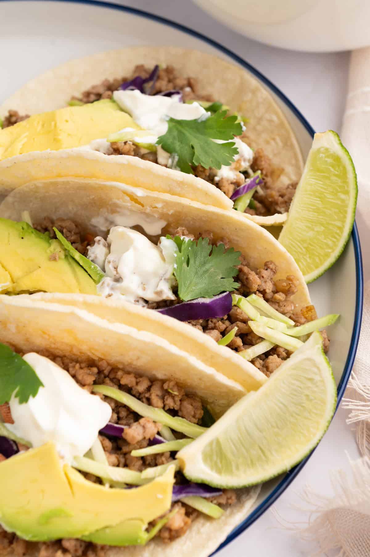 Two ground pork tacos with avocado, sour cream, cilantro, and lime wedges on a plate.