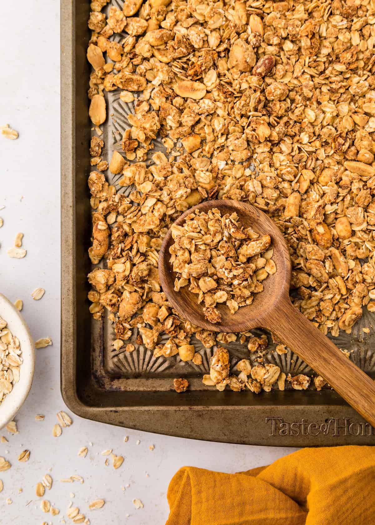 A wooden spoon scoops homemade peanut butter granola on a baking sheet, oats scattered nearby.