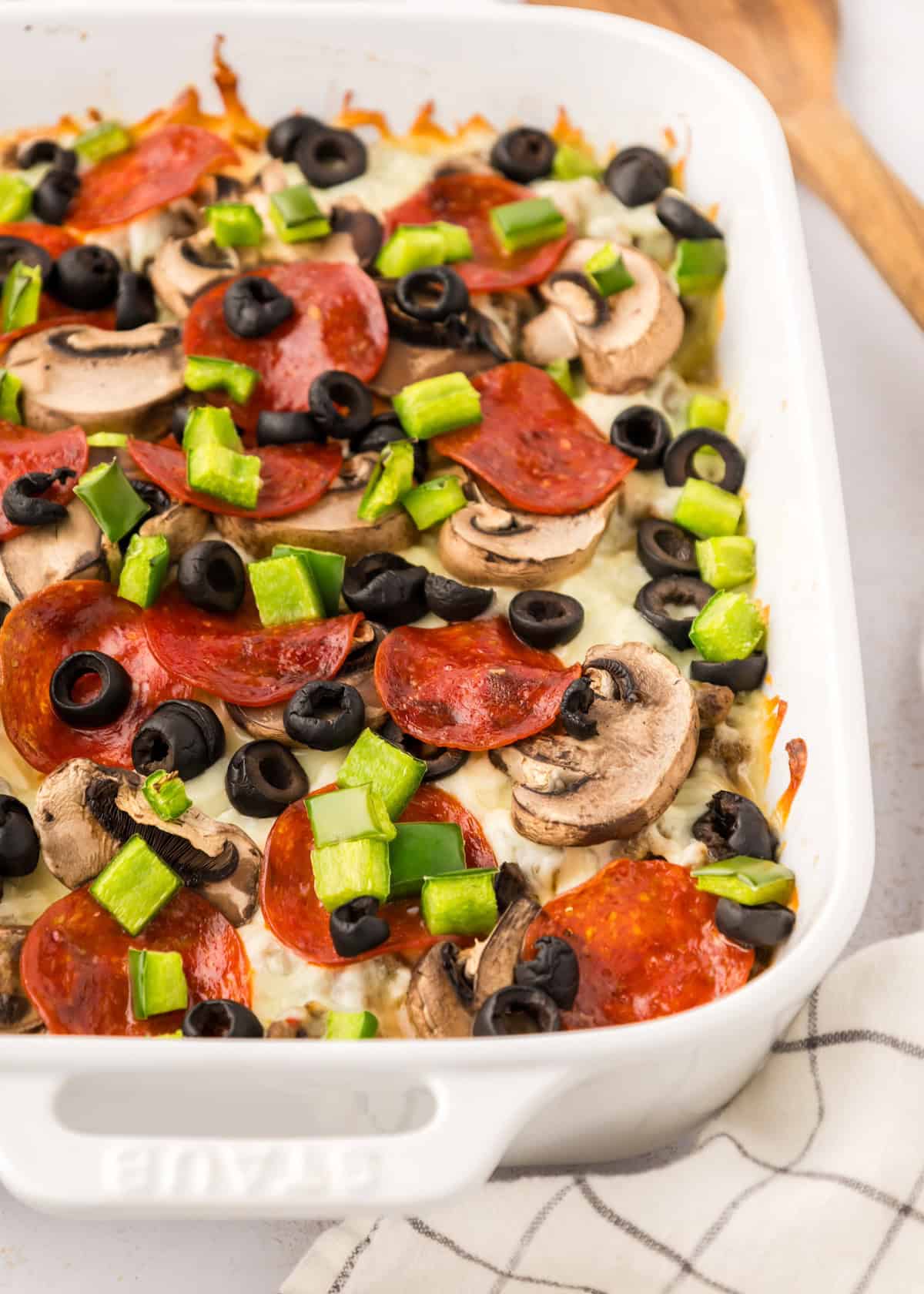 Pizza Casserole topped with pepperoni, mushrooms, green peppers, and black olives in a white dish.