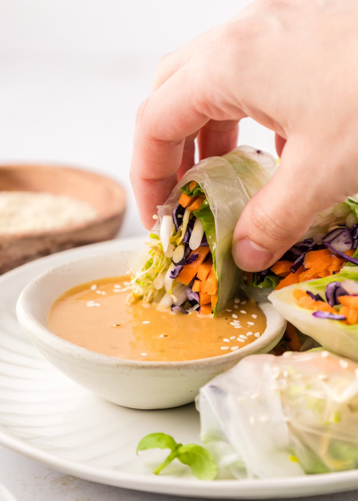 A hand dips a Veggie Spring Roll into peanut sauce topped with sesame seeds.