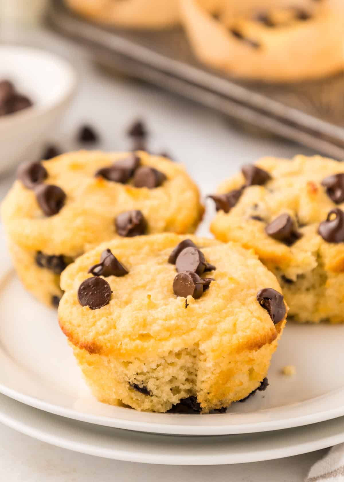 Three healthy chocolate chip muffins on a white plate, with more muffins in the background.