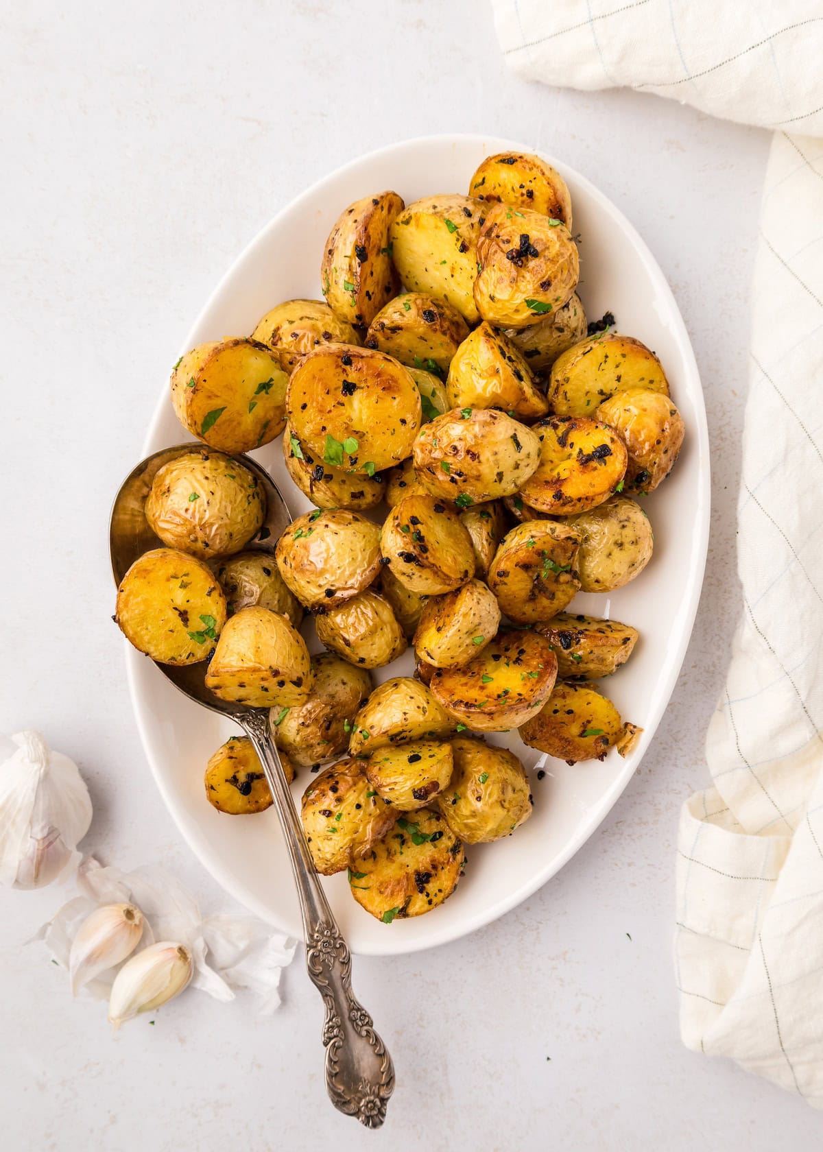 Oval platter of Garlic Roasted Potatoes with herbs, a serving spoon, and fresh garlic cloves.