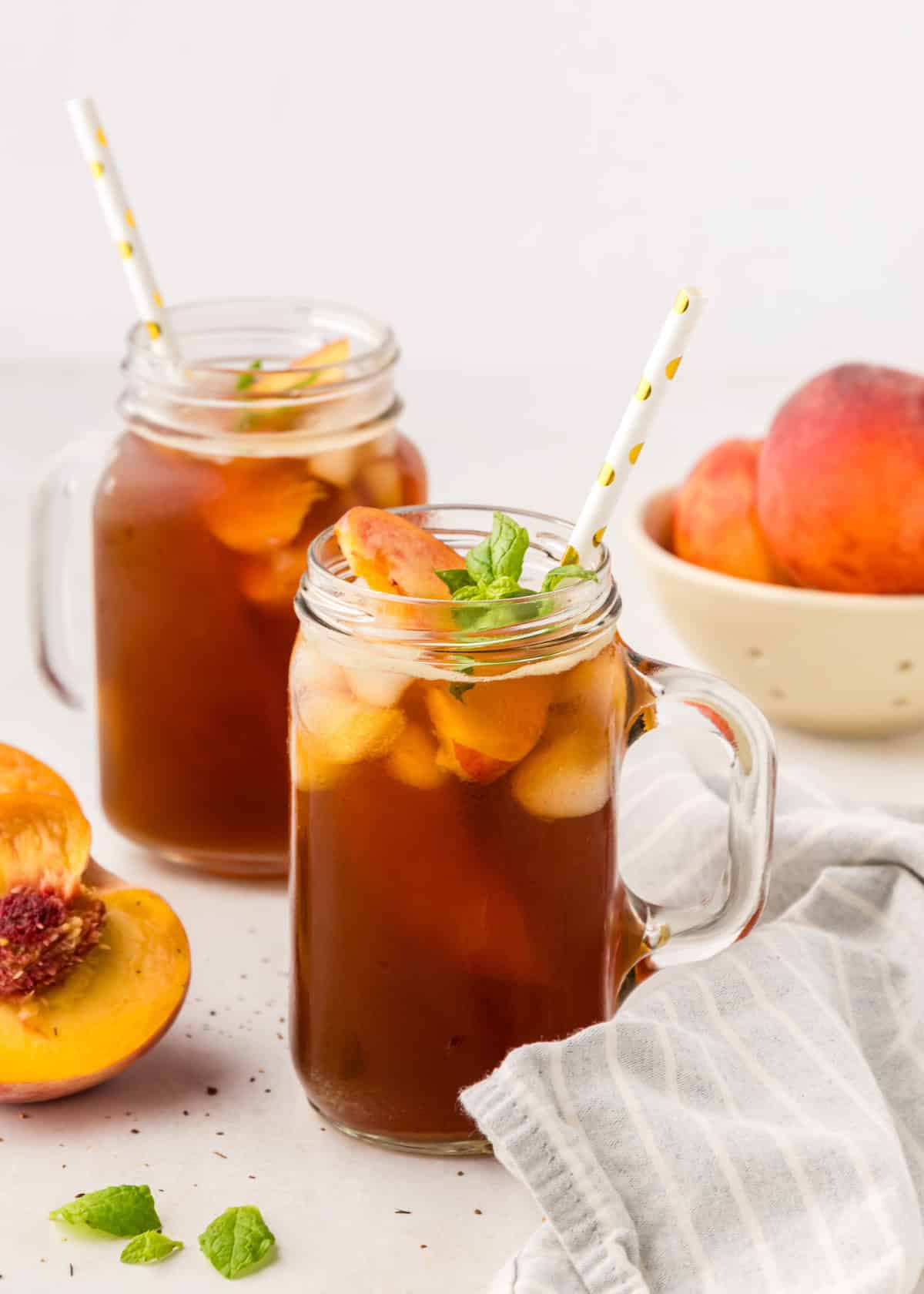 Two mason jars of peach iced tea with straws, fresh peach slices, and mint on a white table.