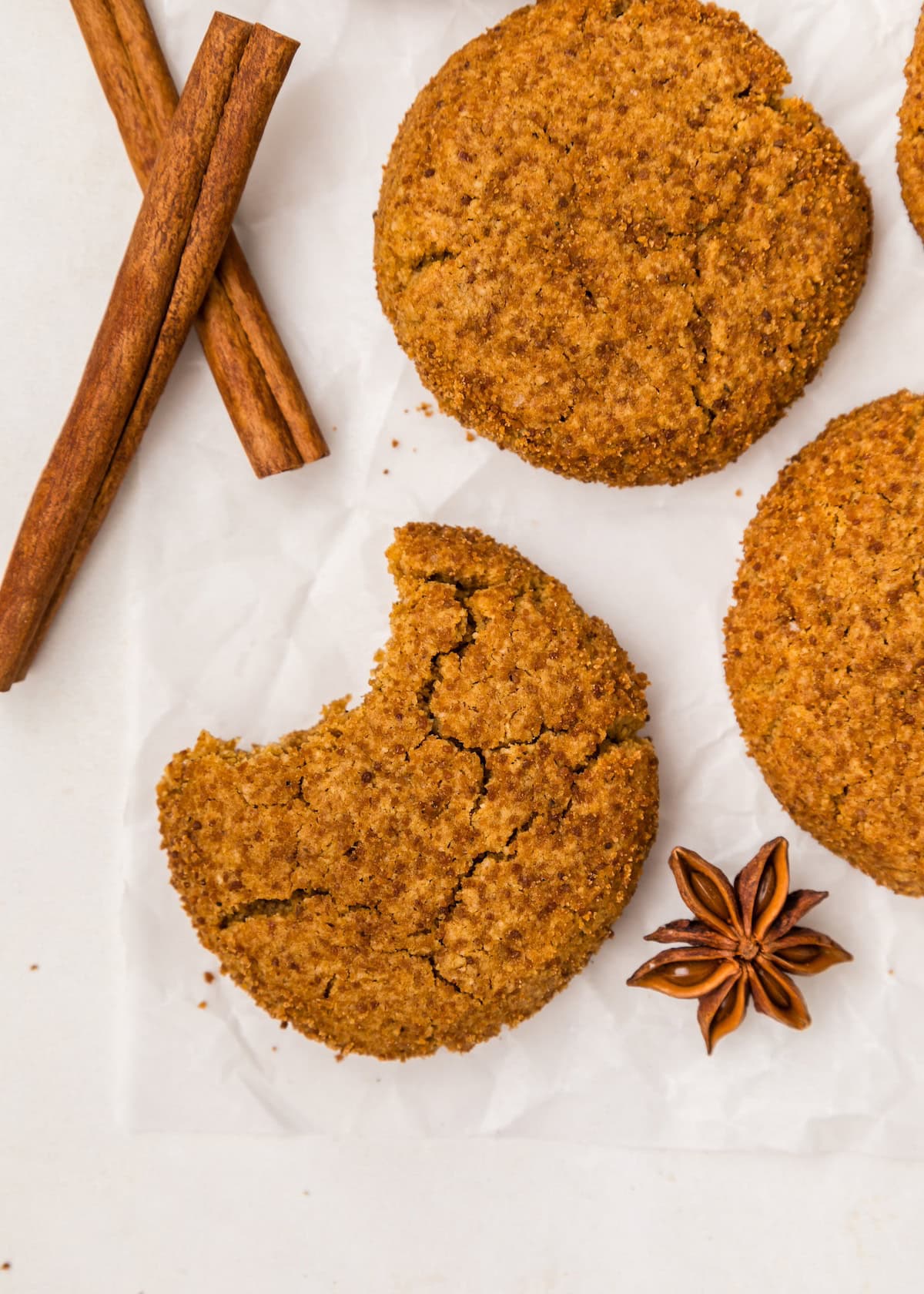 Four Gluten-Free Molasses Cookies, one with a bite, cinnamon sticks, and star anise on parchment.