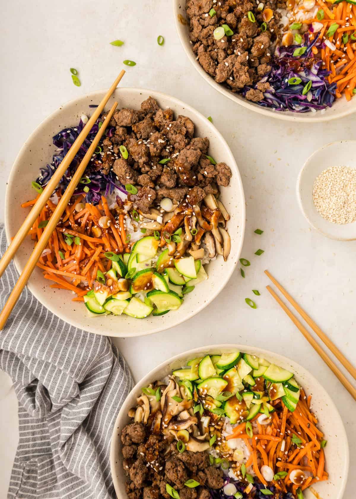 Three korean beef bowls with shredded carrots, zucchini, mushrooms, cabbage, and chopsticks.