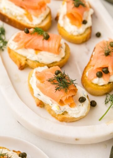 Smoked salmon crostini with cream cheese, dill, and capers served on a white platter.