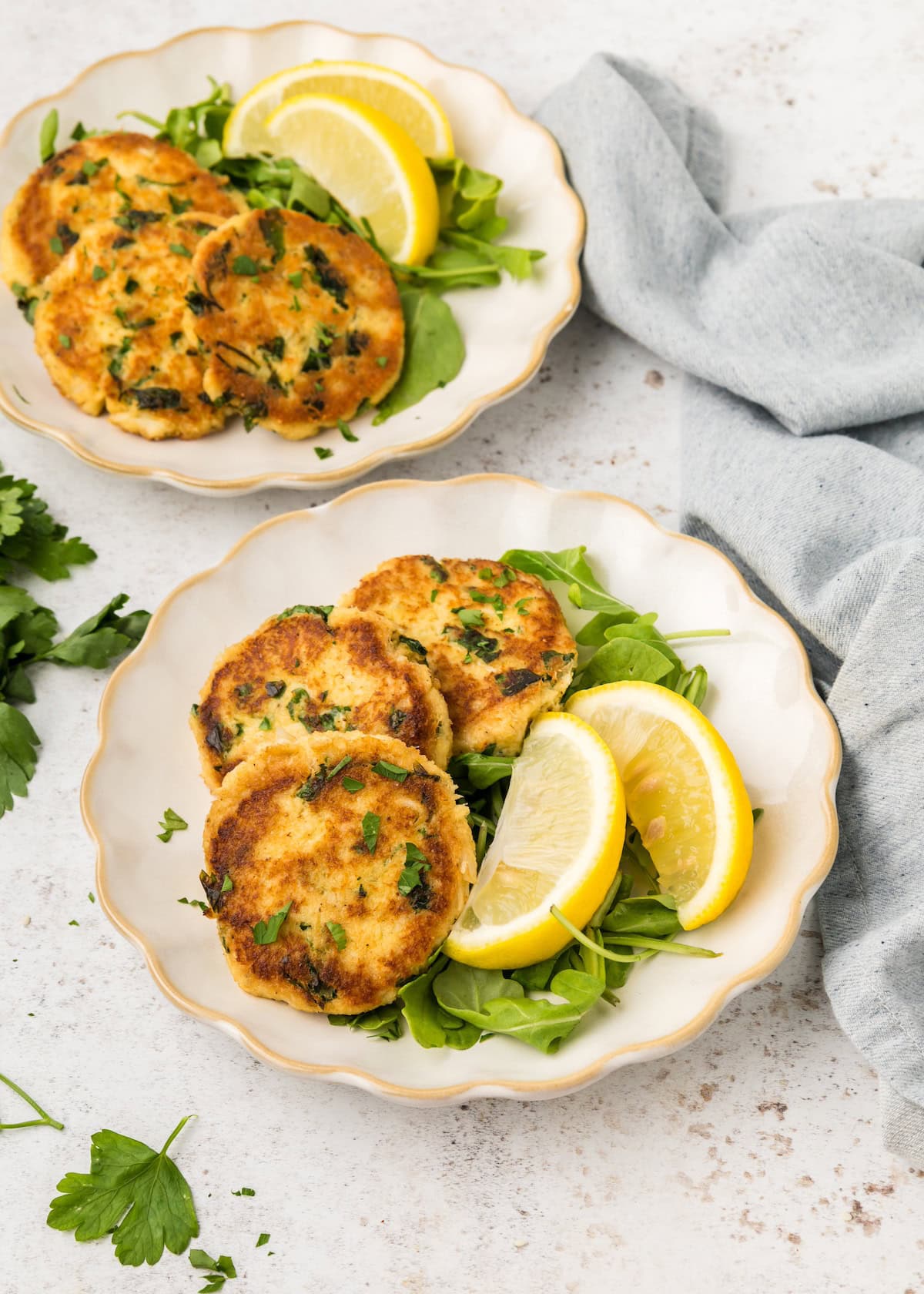 Two plates with gluten-free crab cakes, lemon wedges, and greens on a light surface with a gray napkin.