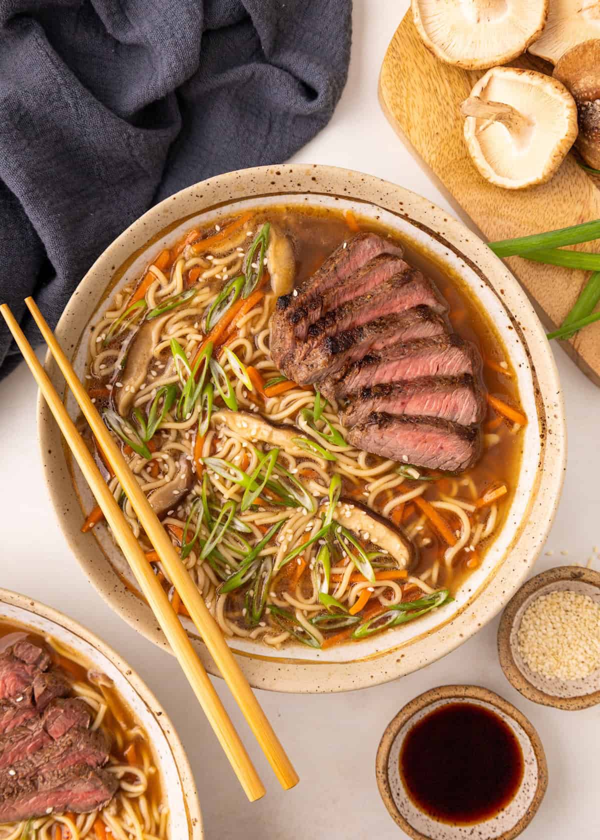Bowl of beef ramen with sliced steak, mushrooms, green onions, and chopsticks on the side.