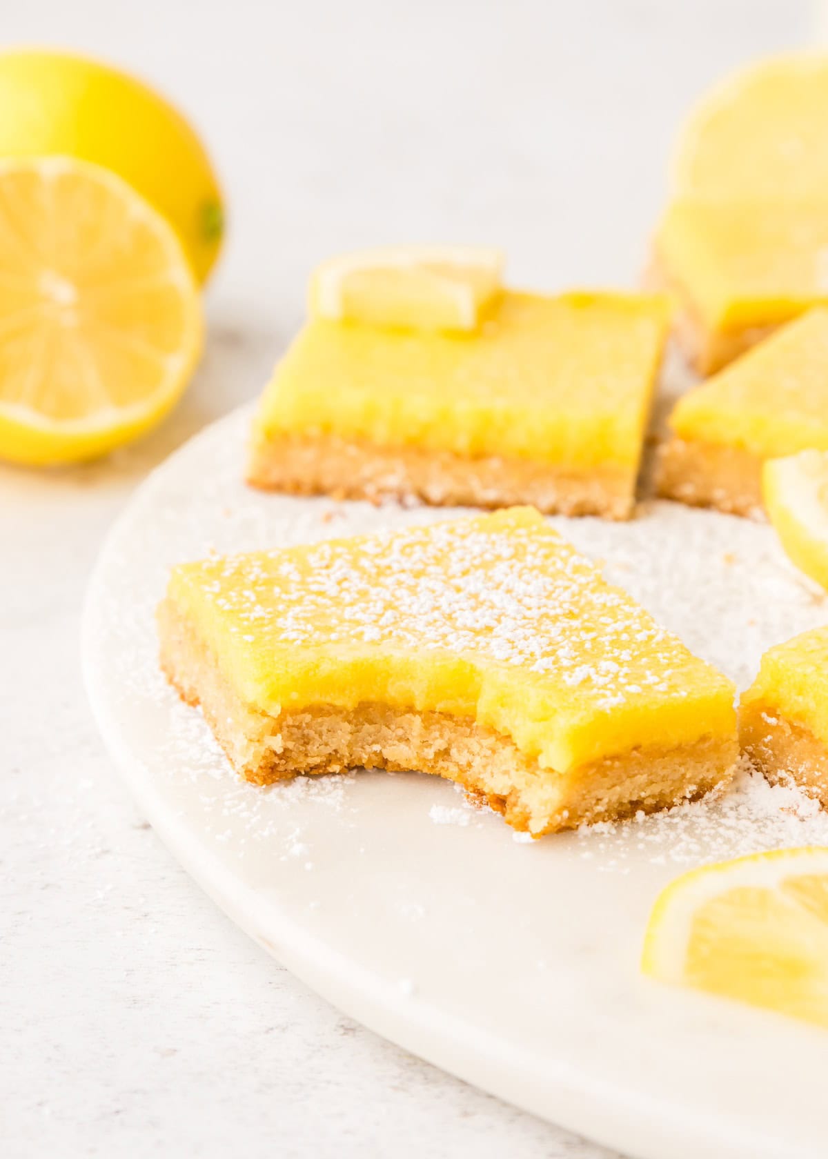 Lemon Bars on a plate, dusted with powdered sugar and lemon slices, one with a bite taken.