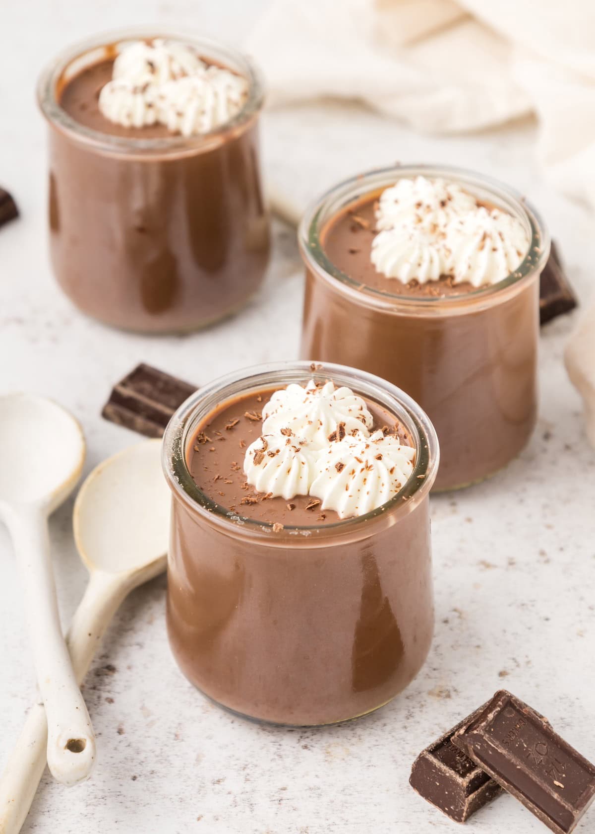 Three jars of Chocolate Pudding topped with whipped cream, chocolate pieces, and spoons nearby.