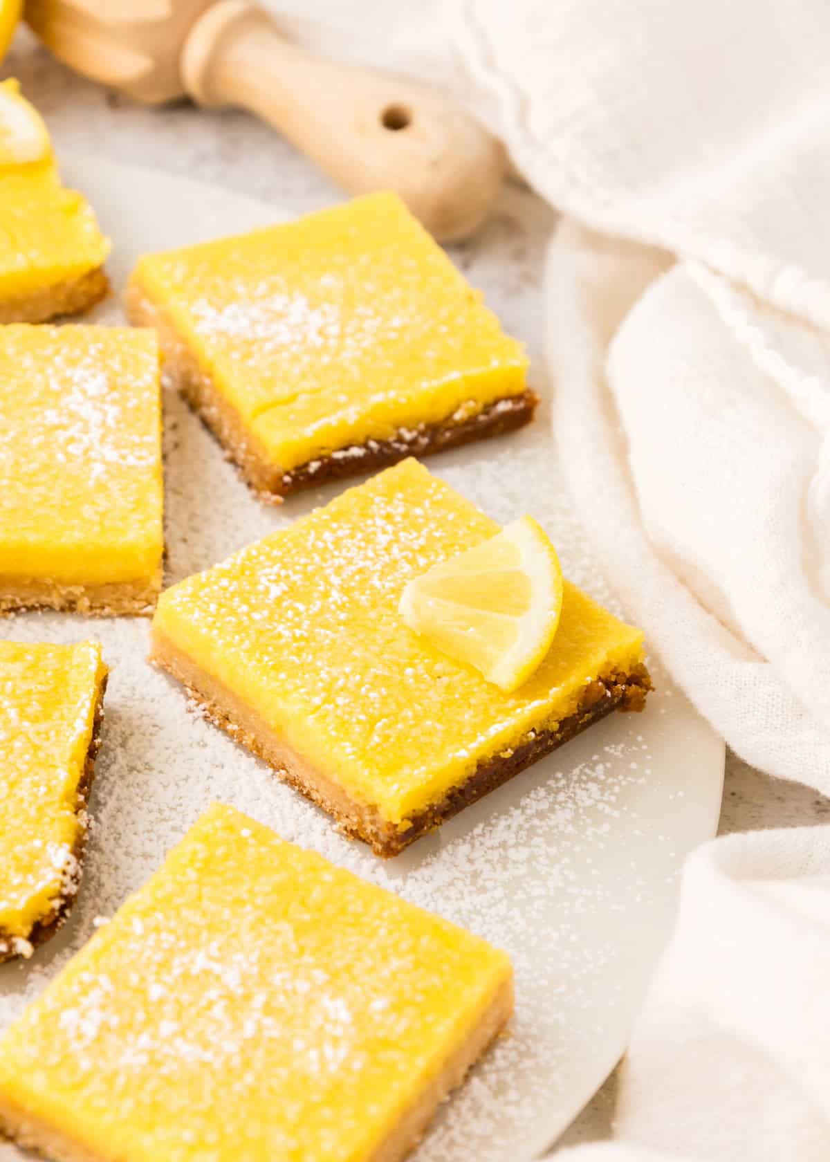 Lemon Bars dusted with powdered sugar, one topped with a lemon slice, on a white plate.