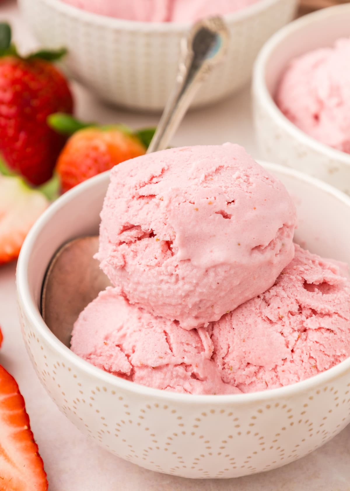Two scoops of strawberry frozen yogurt in a bowl, surrounded by fresh strawberries and a spoon.