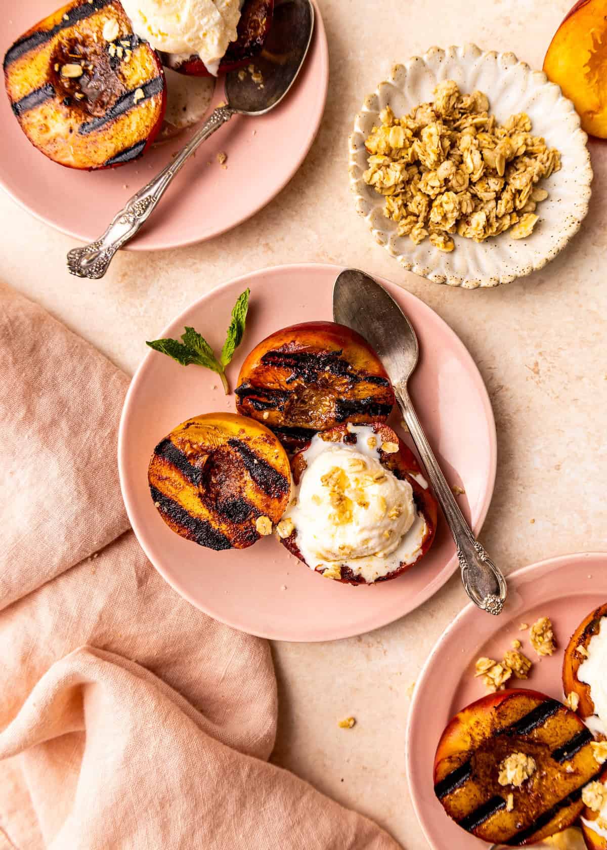 Honey Grilled Peaches with ice cream and granola on pink plates, mint garnish, and a spoon.