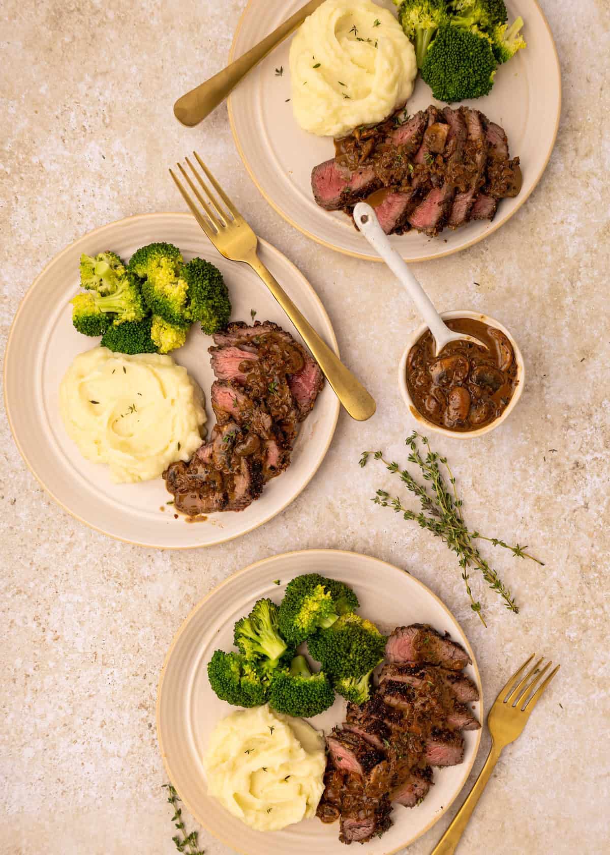 Two plates of steak with mushroom sauce, mashed potatoes, and broccoli on a light surface.