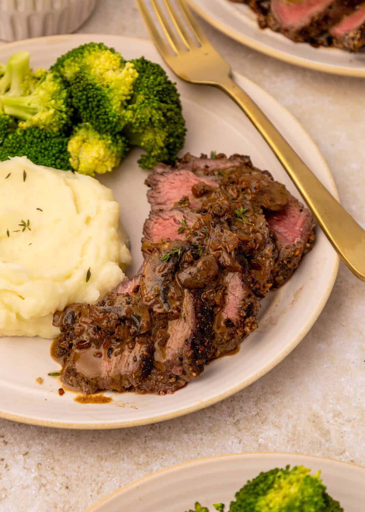 Steak with mushroom sauce, mashed potatoes, and broccoli served on a plate with a gold fork.