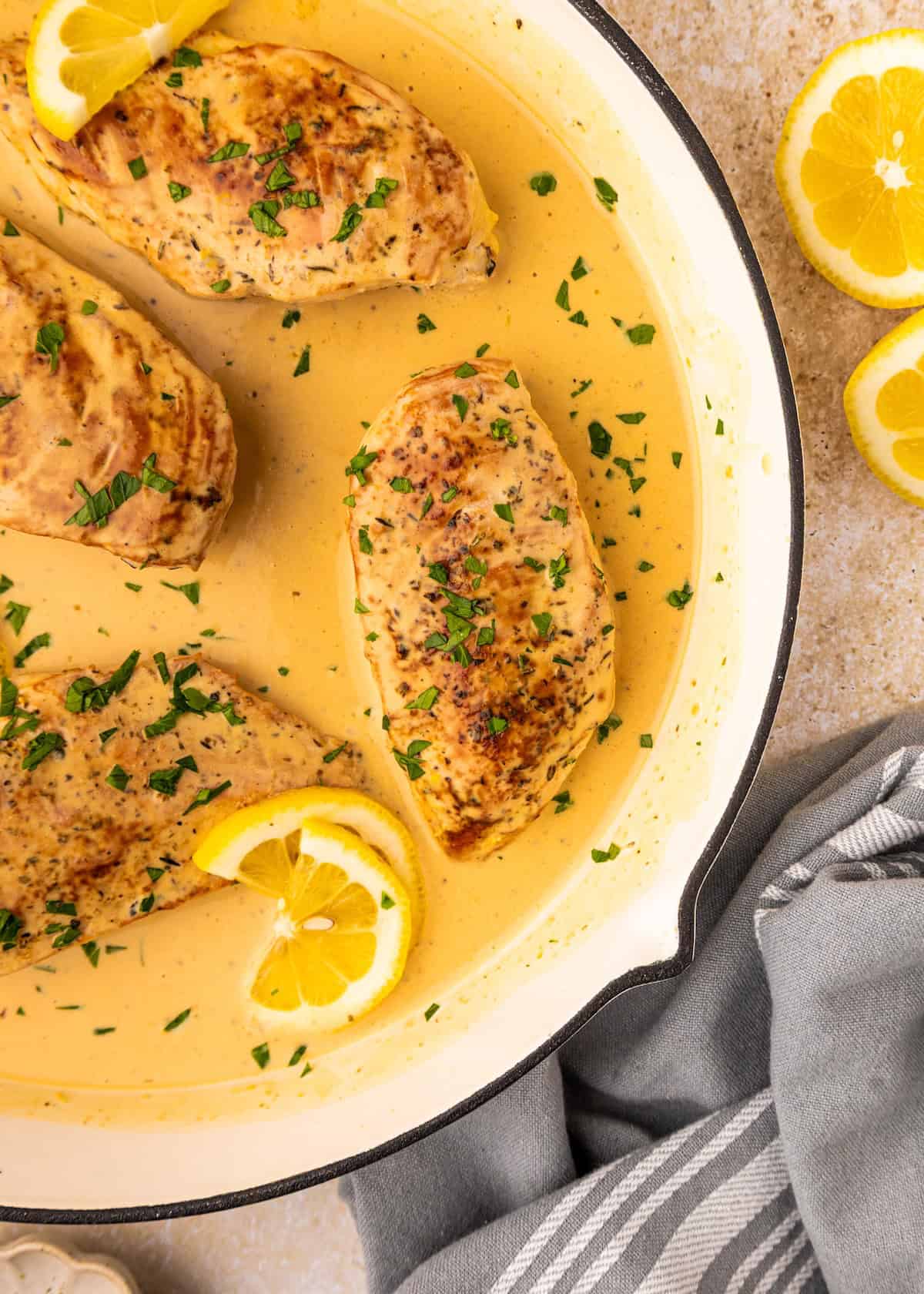 Creamy Lemon Chicken breasts in a pan with herbs, lemon slices, and a light sauce, overhead view.