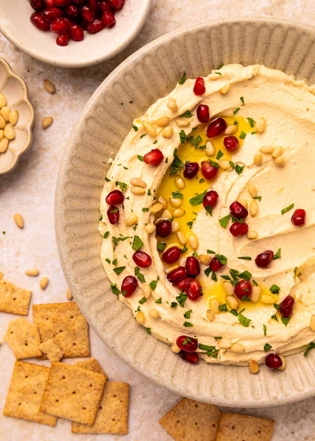 Toasted Pine Nut Hummus with pomegranate seeds, herbs, olive oil, and crackers nearby.
