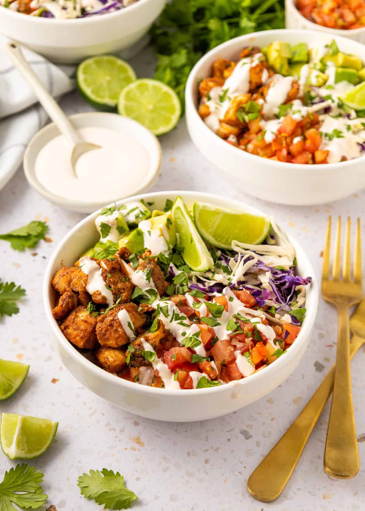 Fish Taco Bowls with seasoned chicken, veggies, lime wedges, creamy sauce, and gold utensils nearby.