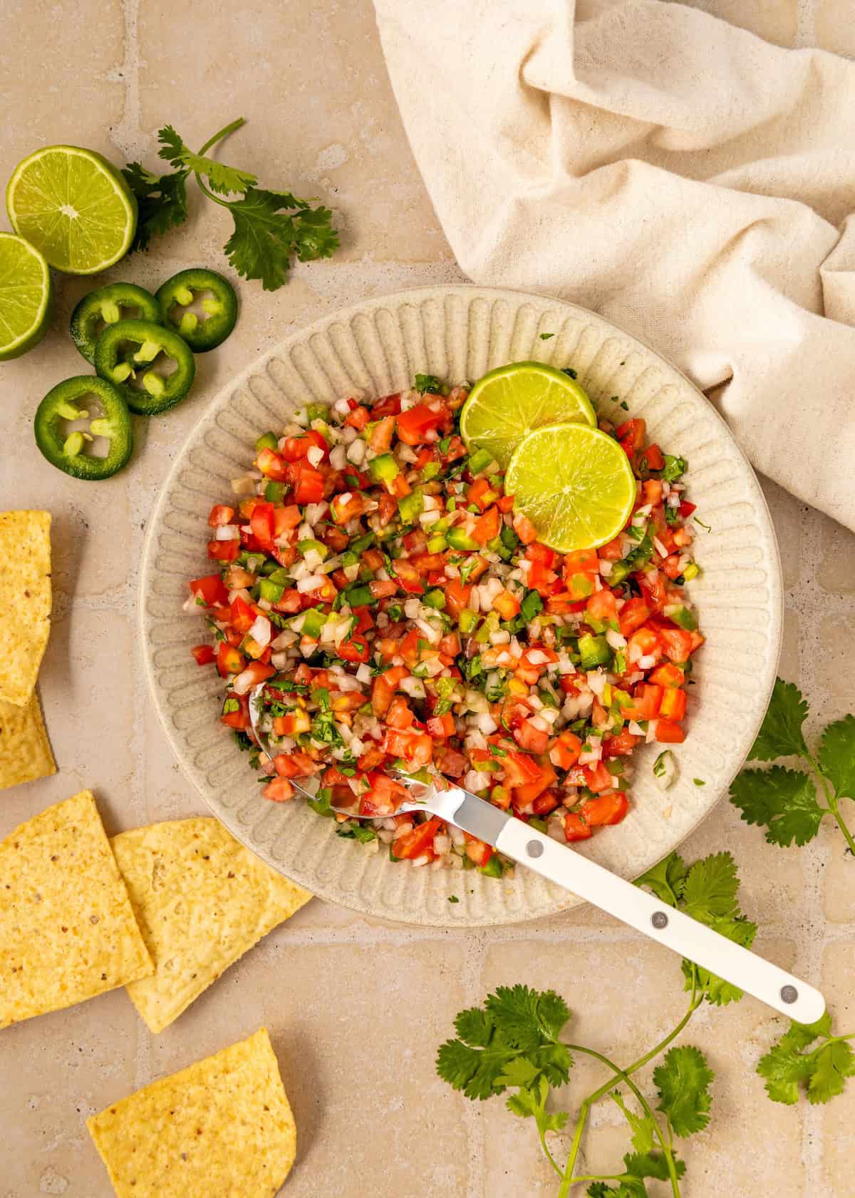 A bowl of fresh Pico de Gallo with lime slices, tortilla chips, jalapeño, and cilantro on the side.
