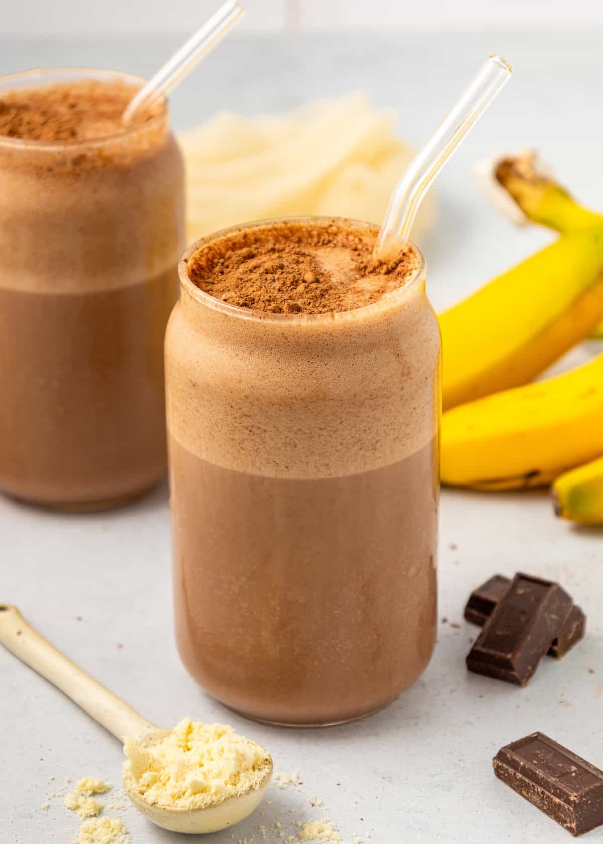 Two glasses of Chocolate Protein Shake with glass straws, bananas, and chocolate nearby.