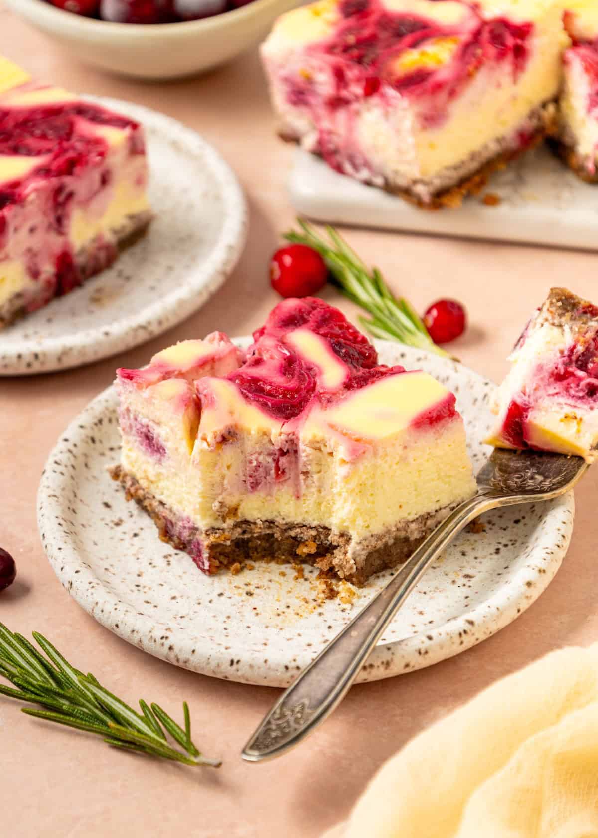A Cranberry Cheesecake Bars slice on a speckled plate with rosemary garnish and a fork.