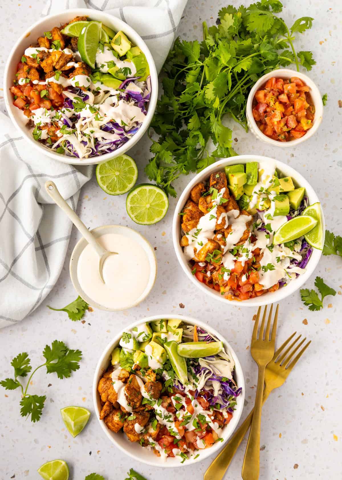 Three Fish Taco Bowls with veggies, avocado, sauce, and lime, surrounded by cilantro and salsa.