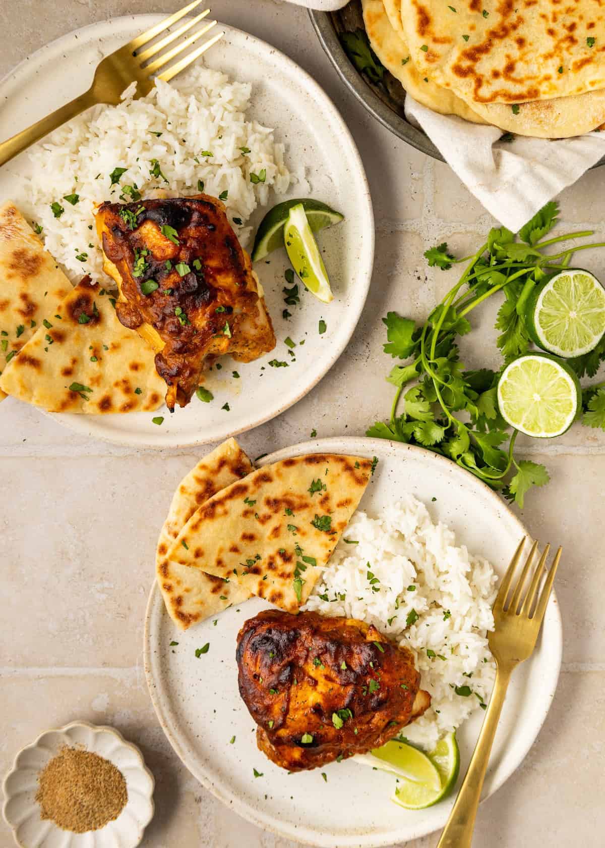 Two plates with rice, naan bread, tandoori chicken, lime wedges, and fresh herbs on a table.