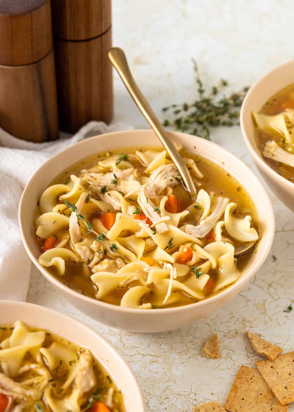 Bowl of turkey noodle soup with carrots, herbs, and a spoon, surrounded by crackers.