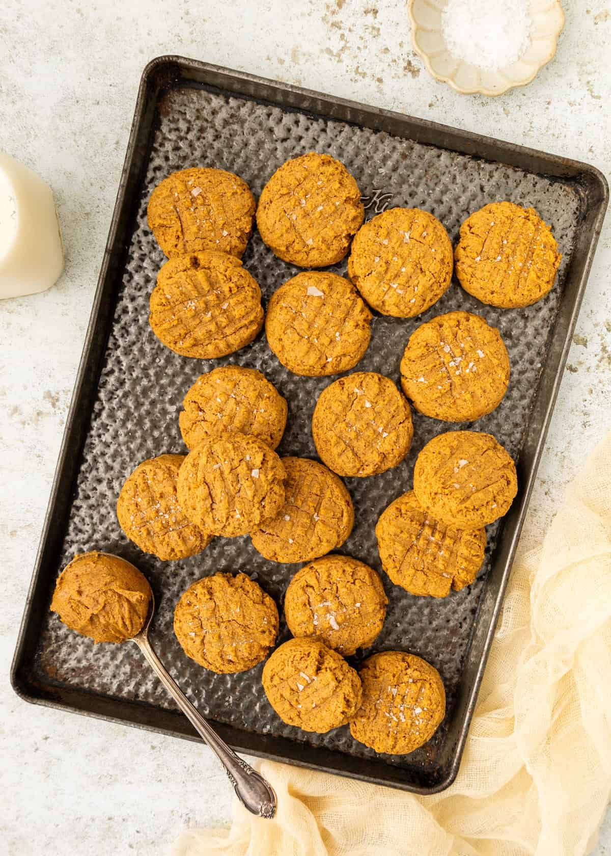 A tray of round almond butter cookies with a spoon and a small bowl of salt nearby.