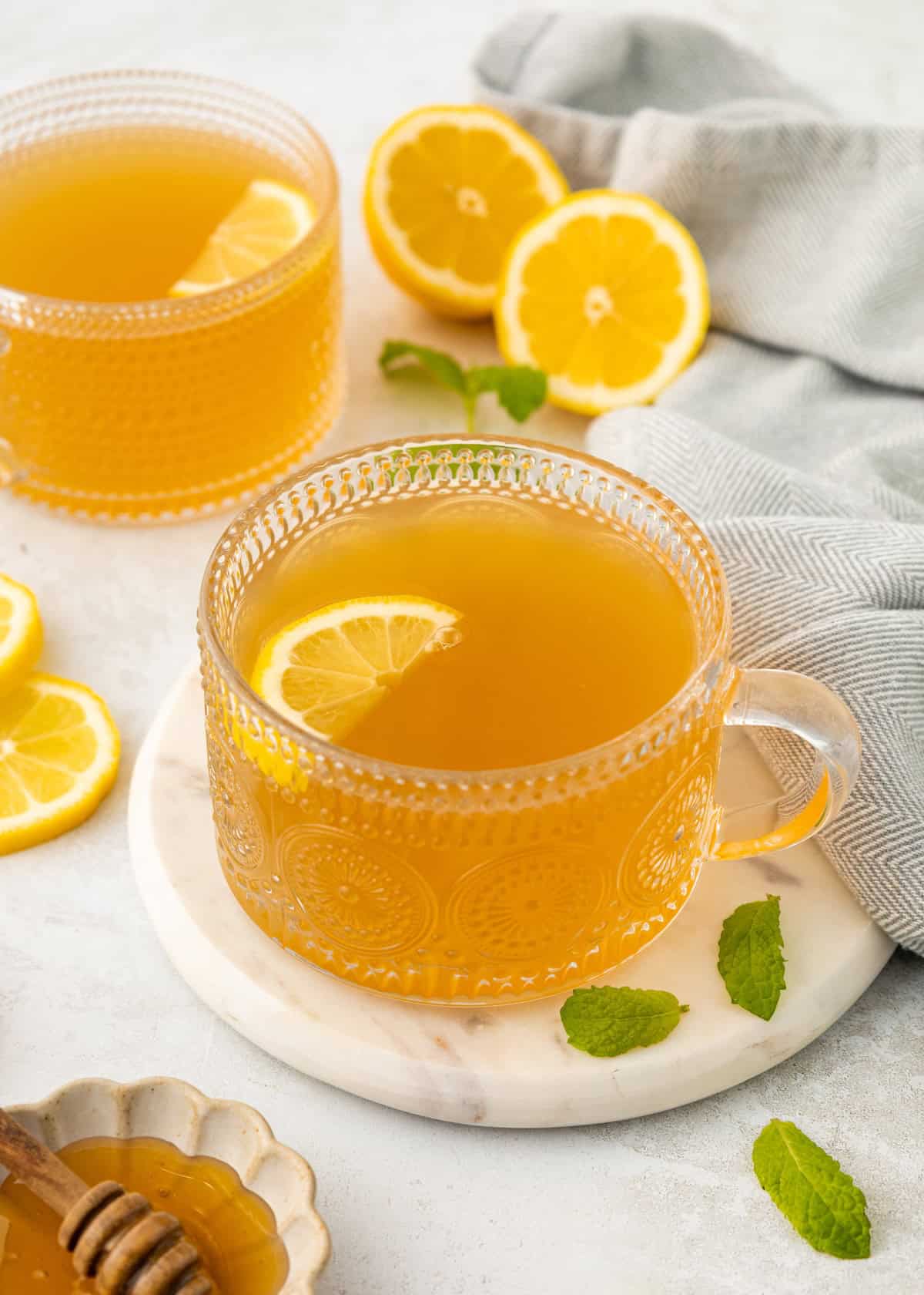 Glass cup of Medicine Ball Tea with lemon slice, fresh mint, and lemon halves on a light surface.